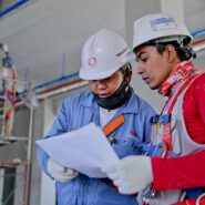 A pair of factory workers wearing protective equipment consult some papers.